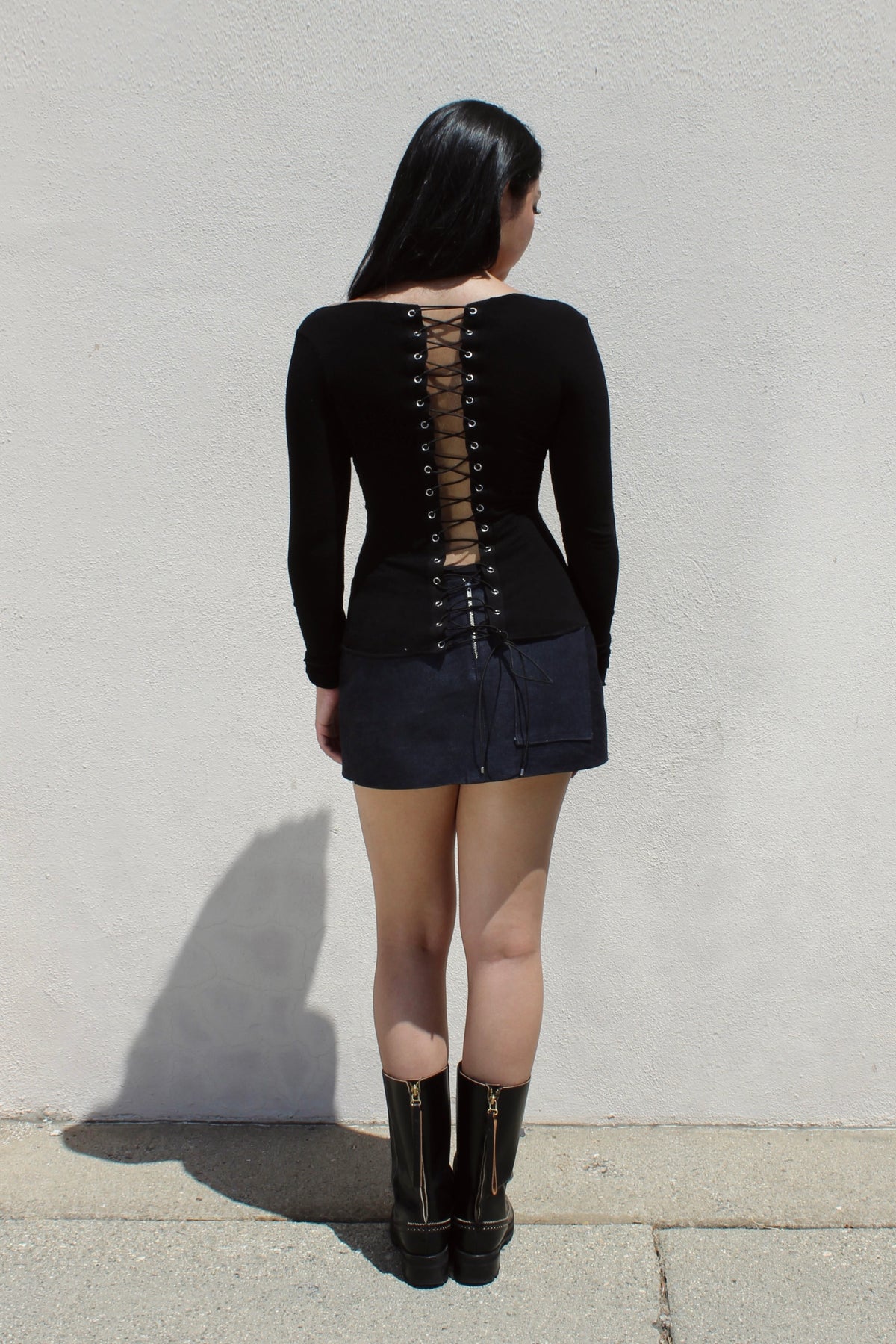 Lace-up back of the Ivy top in black, worn with a denim mini skirt against a white wall