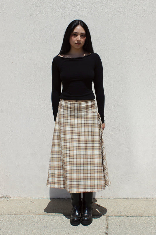 Aster skirt in tan plaid worn with a black long-sleeve top against a white stucco wall