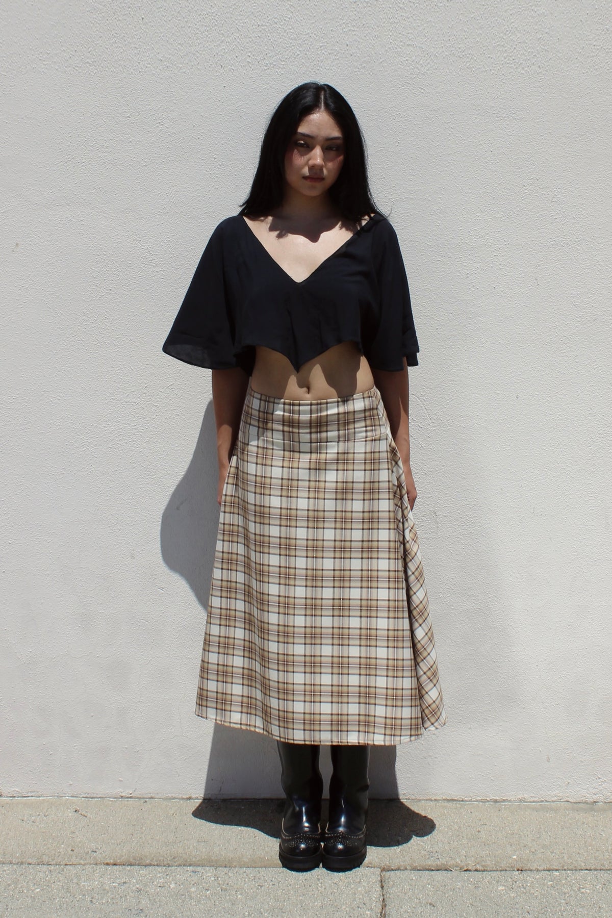 Lotus top in black worn with the Aster skirt in tan plaid against a white stucco wall