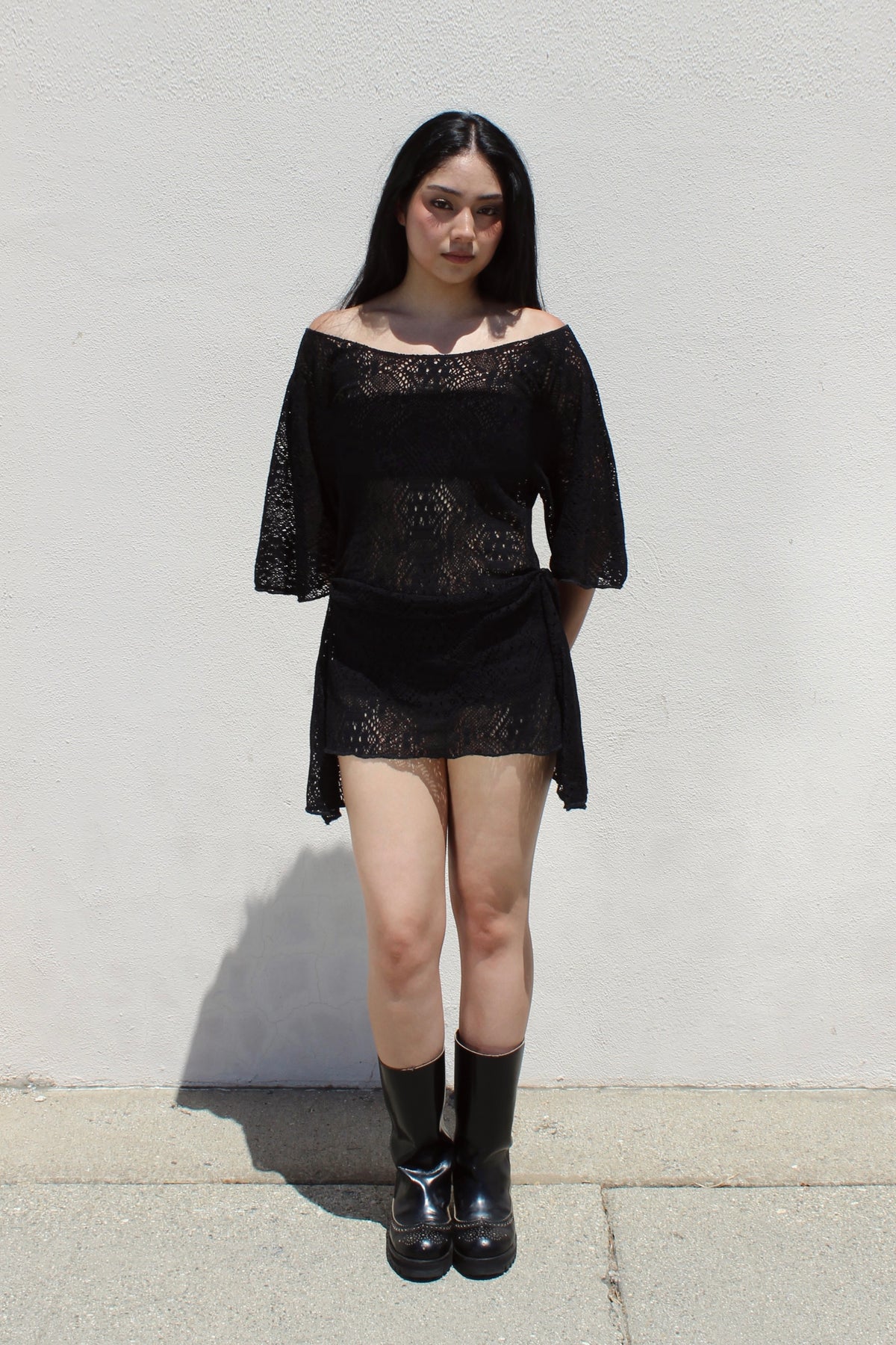 Dahlia dress in black lace worn with tall boots against a white stucco wall